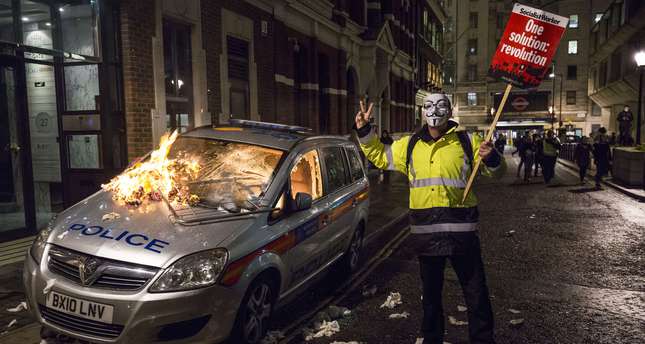 An anti-capitalist protester wearing a Guy Fawkes mask holds a placard as he stands alongside a burning police car during the ,Million Masks March,, organised by the group Anonymous, in London on November 5, 2015. (AFP Photo)