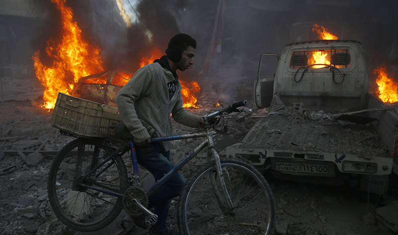 A man carries his bicycle past debris and burning cars following reported airstrikes in the town of Hamouria, Syria (AFP Photo)
