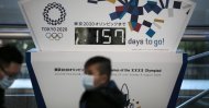 People wearing masks sit in front of a countdown clock for the 2020 Olympics in Tokyo, Feb. 18, 2020. (AP Photo)