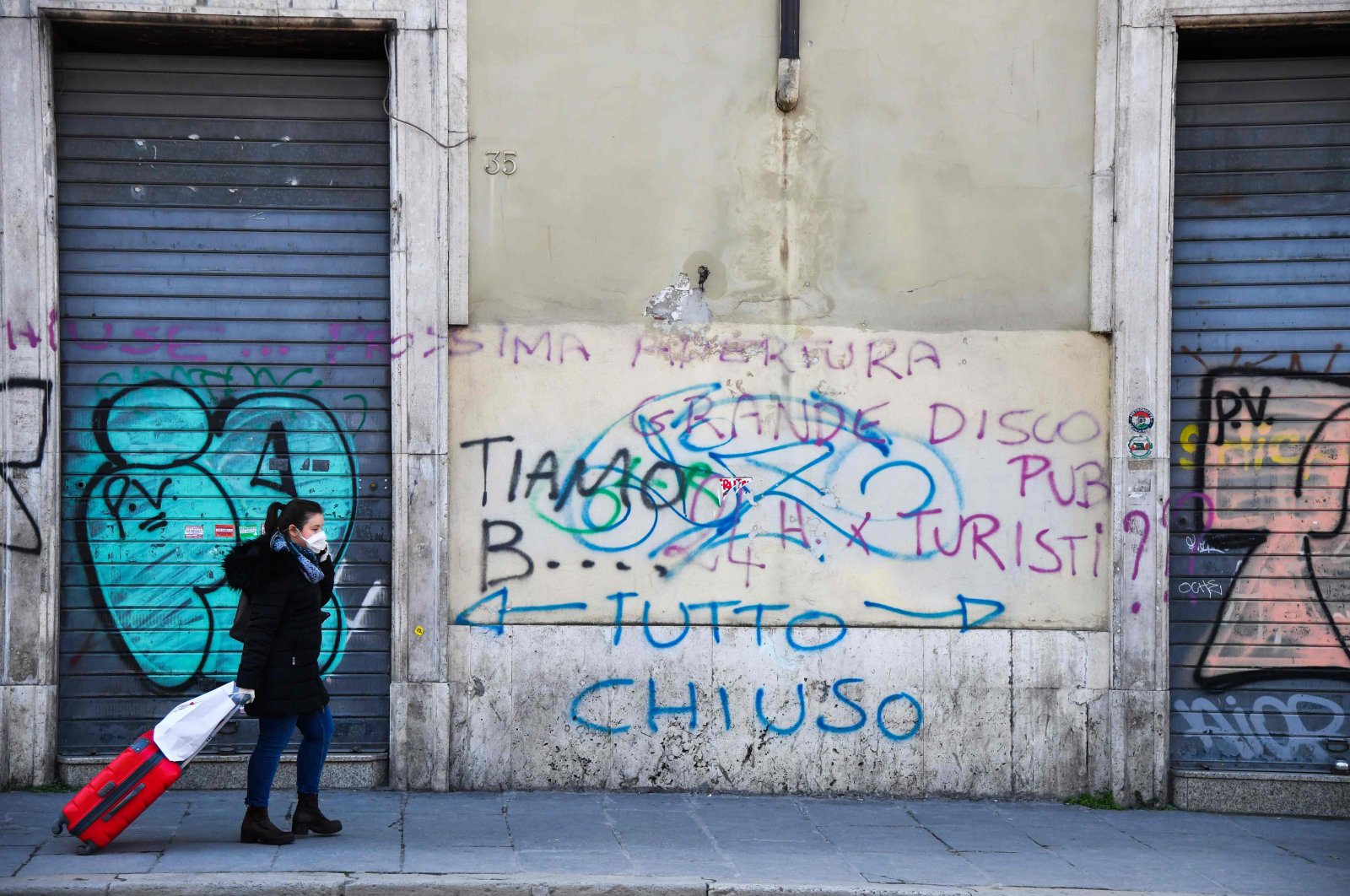 A commuter wearing a protective mask walks past closed shops and an inscription on the wall reading "All Closed," Rome, March 11, 2020. (AFP Photo)