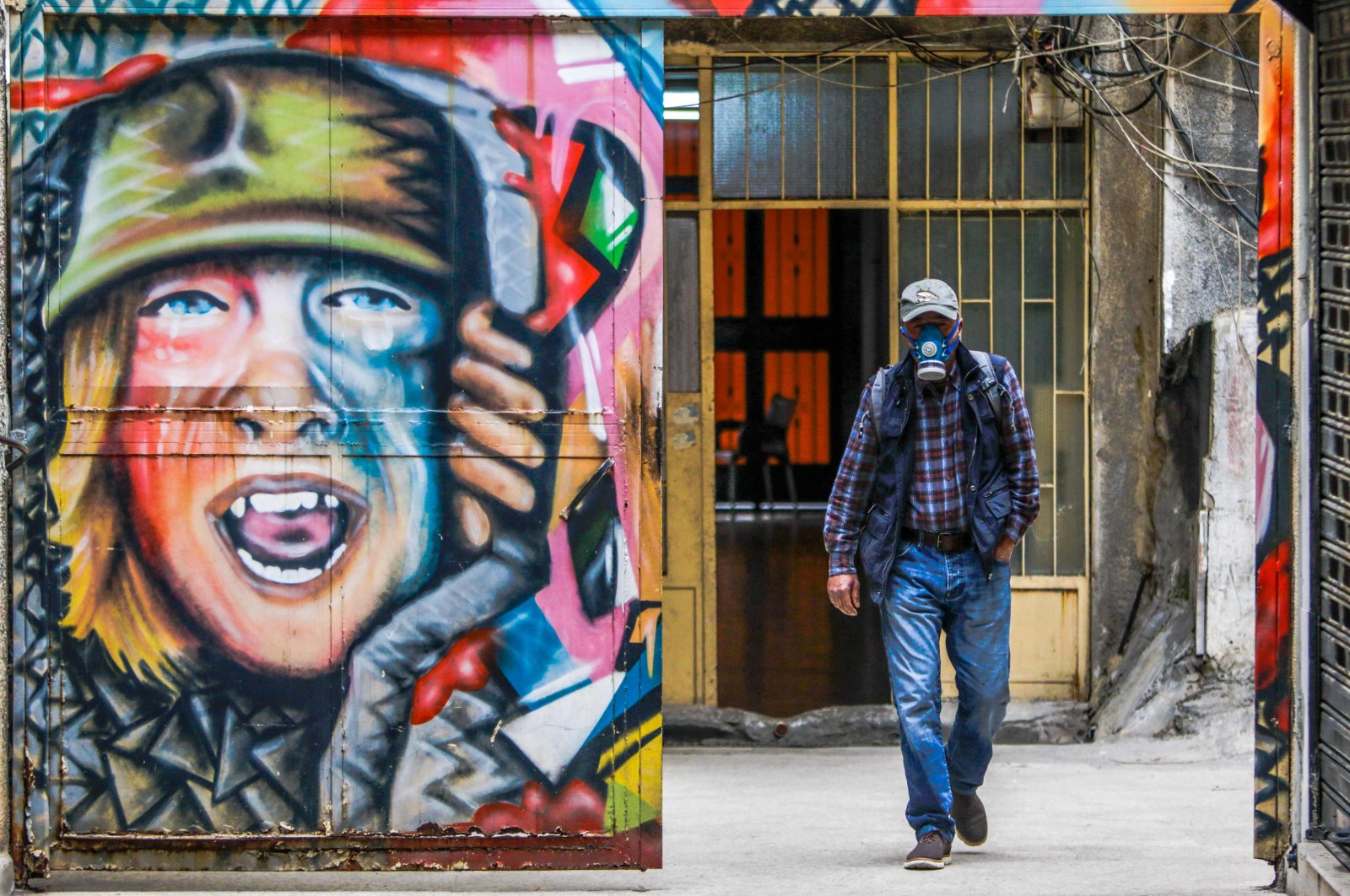 A man, wearing facial gas mask as part of health safety measures related to the spread of COVID-19, walks past a graffiti in the northern port city of Tripoli, Monday, March 16, 2020. (AFP Photo)