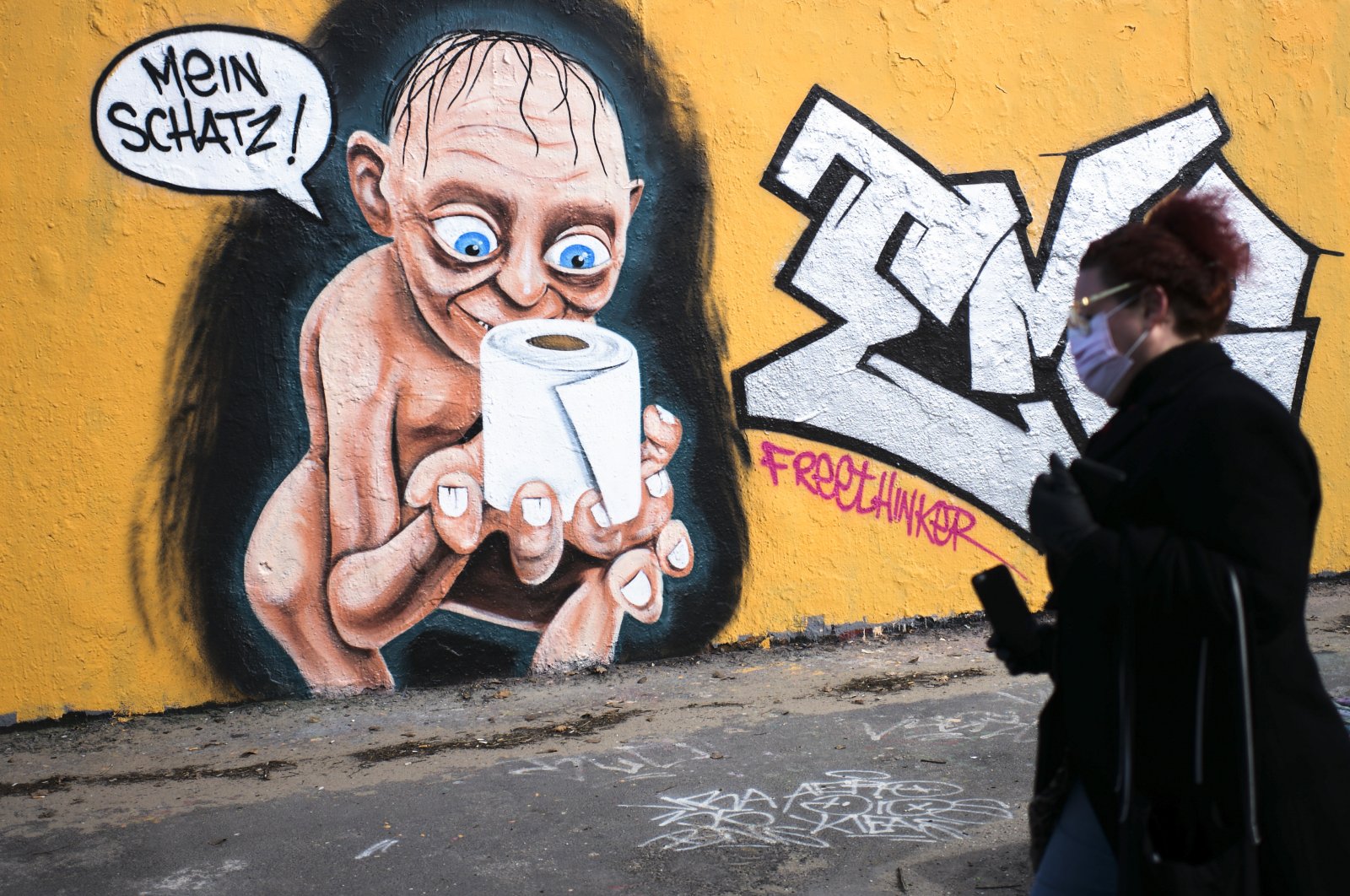 A woman walks in front of graffiti depicting the character of Gollum from "Lord of the Rings," holding a roll of toilet paper and saying "My precious," in the Mauerpark public park in the Prenzlauer Berg district of Berlin, Germany, Saturday, March 21, 2020. (AP Photo)