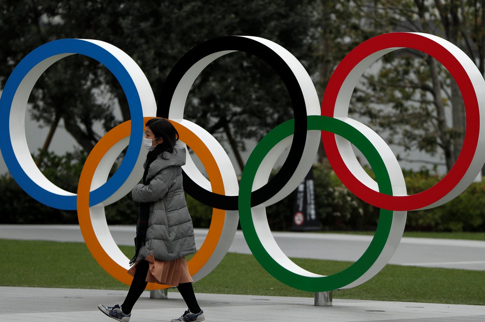 A woman wearing a protective face mask walks past the Olympic rings in front of the Japan Olympics Museum, in Tokyo, Japan, Monday, March 30, 2020. (Reuters Photo)