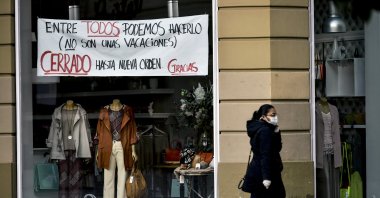 A woman wearing a face mask walks in front of a store with a banner reading, "Together we can do it. It's not the holidays. Closed until further notice. Thank you," Pamplona, Spain, April 8, 2020. (AP Photo)