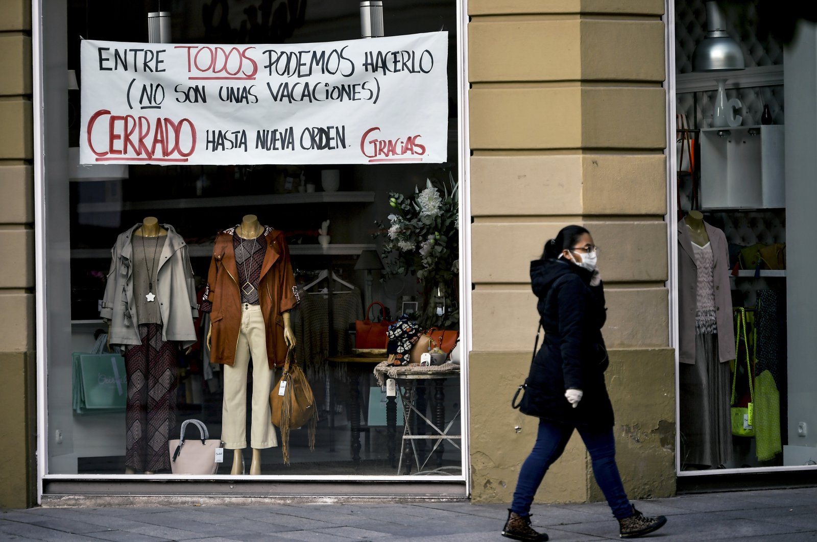 A woman wearing a face mask walks in front of a store with a banner reading, "Together we can do it. It's not the holidays. Closed until further notice. Thank you," Pamplona, Spain, April 8, 2020. (AP Photo)