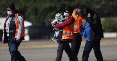 An immigration worker, wearing an orange jacket and a mask as a precaution against the coronavirus, carries a young Guatemalan migrant who was deported from the U.S., followed by another deportee, at La Aurora International Airport, Guatemala City, March 12, 2020. (AP Photo)