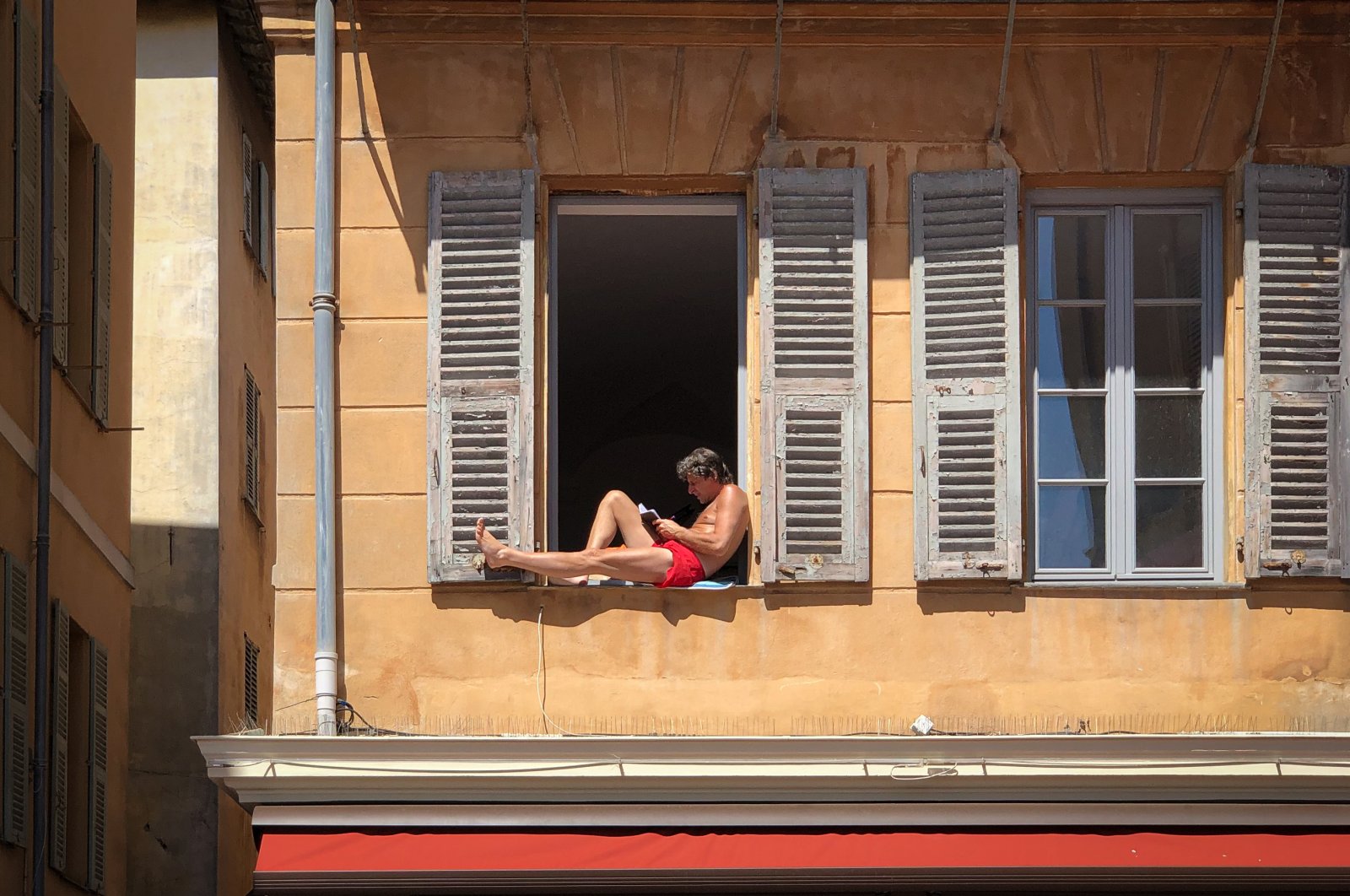 A man alone and confined to his apartment, sunbathing shirtless while reading a book in his window, in old Nice. (Reuters Photo)