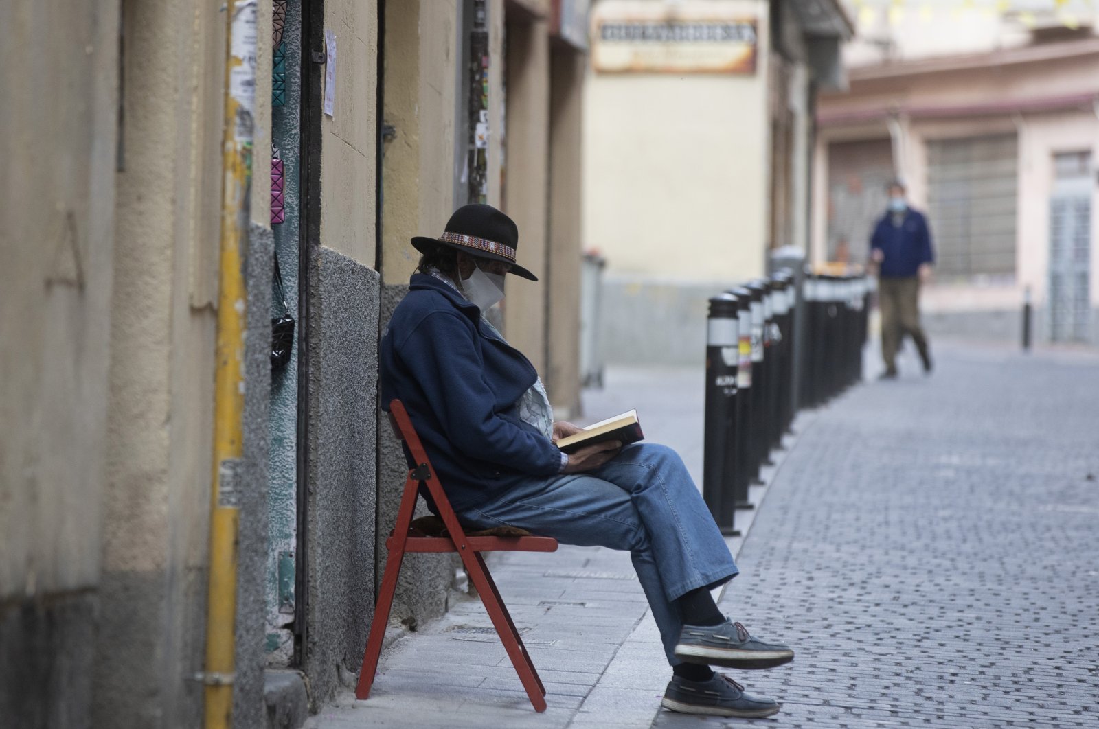 A man wearing a face mask to protect against coronavirus sits and waits for customers outside a small shop while reading a book in Madrid, Spain, Saturday, May 16, 2020.  (AP Photo)