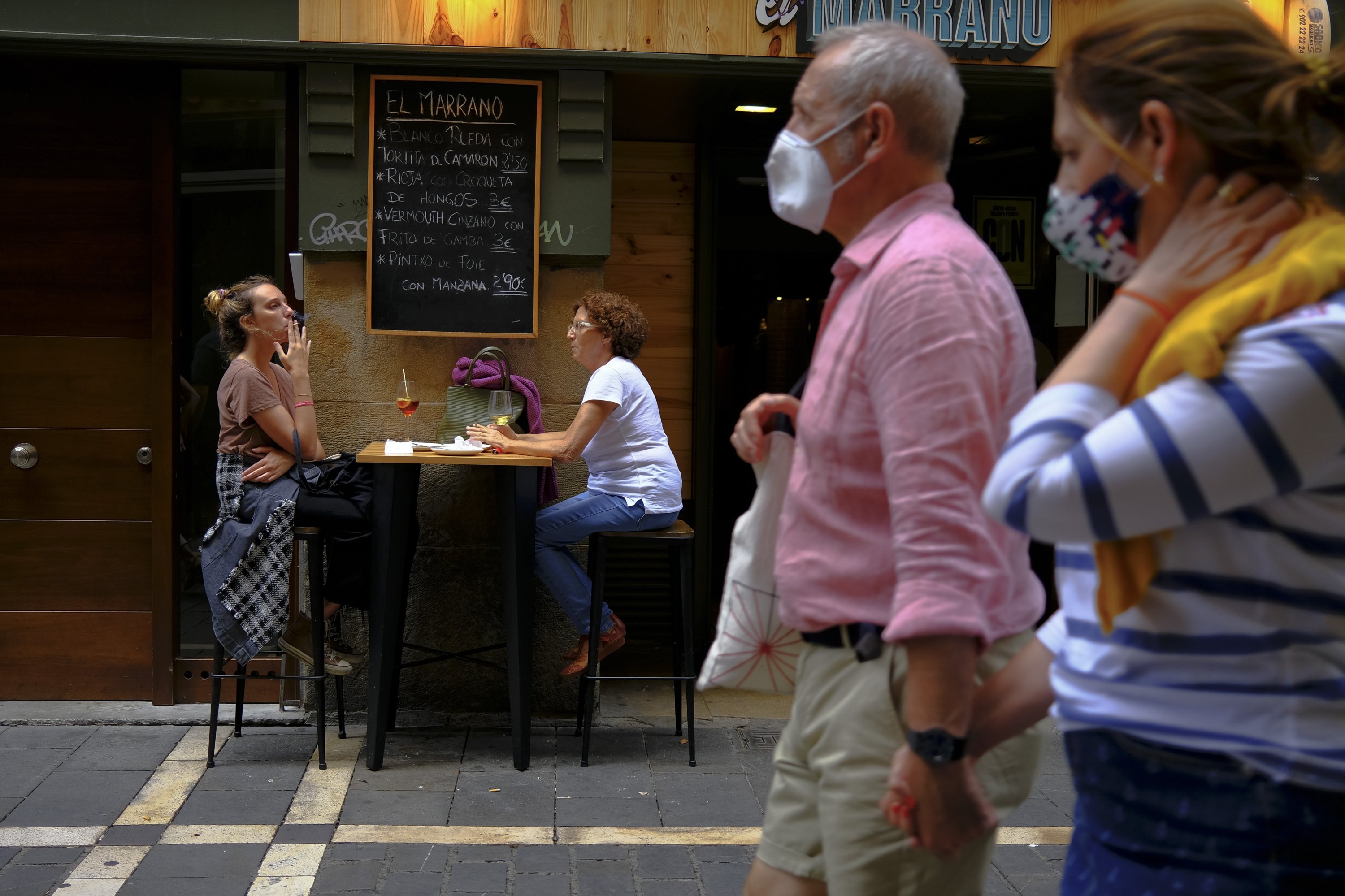 Two women smoke cigarettes in a bar as pedestrians wearing face masks pass by in Pamplona, ​​northern Spain, on Aug.17, 2020. (AP Photo)