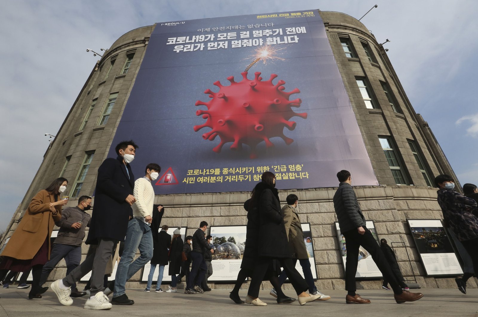 People wearing masks as a precaution against the coronavirus walk under a banner emphasizing an enhanced social distancing campaign in front of Seoul City Hall in Seoul, South Korea, on Nov. 25, 2020. (AP Photo)