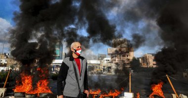 A man clad in a mask depicting the Lebanese flag stands next to flaming tires at a makeshift roadblock set up by anti-government demonstrators in the area of Dora on the northern outskirts of Lebanon's capital Beirut on March 8, 2021. (AFP Photo)