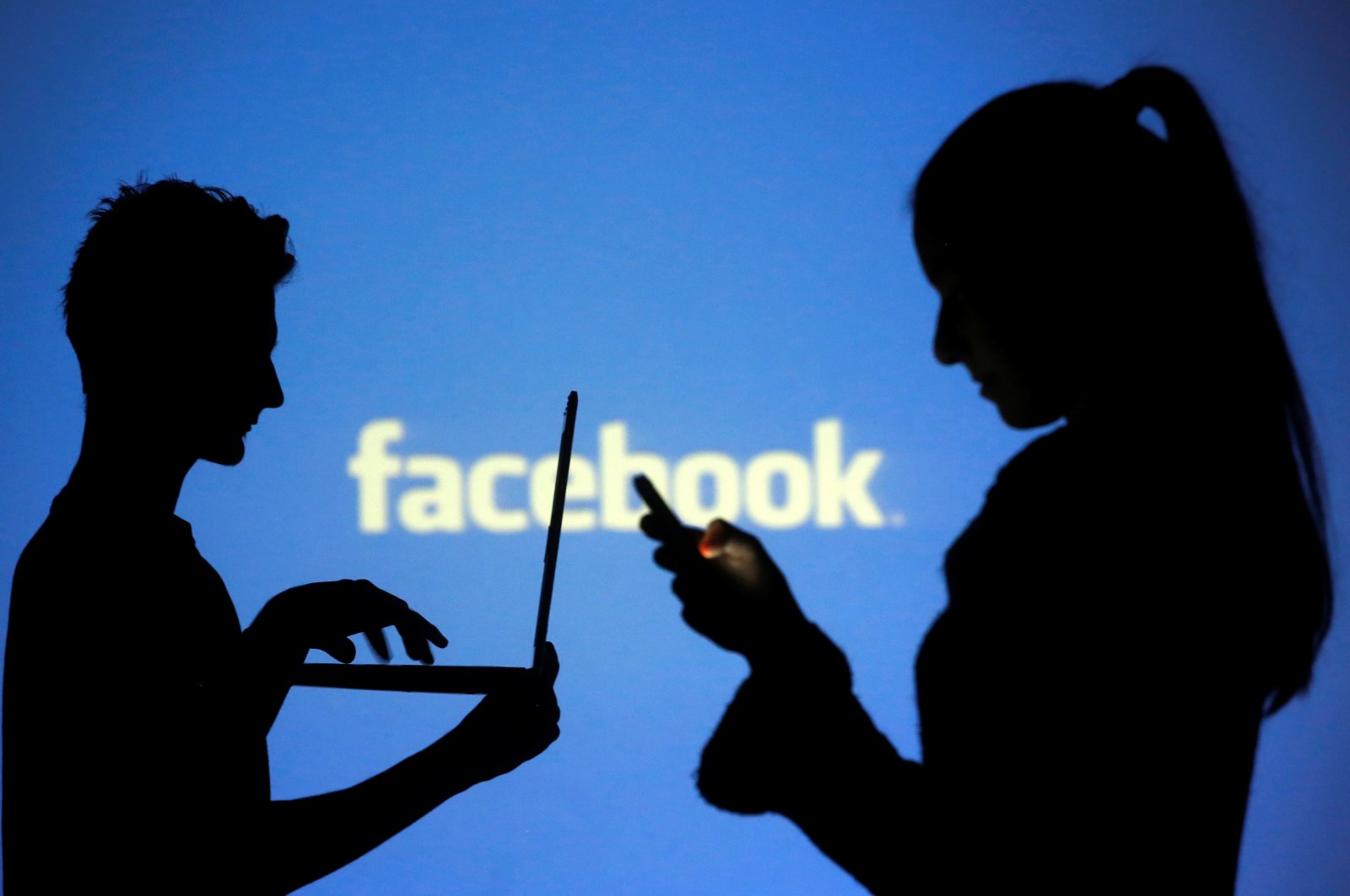 People are silhouetted as they pose with laptops in front of a screen projected with a Facebook logo, in this picture illustration, Oct. 29, 2014. (Reuters Photo)