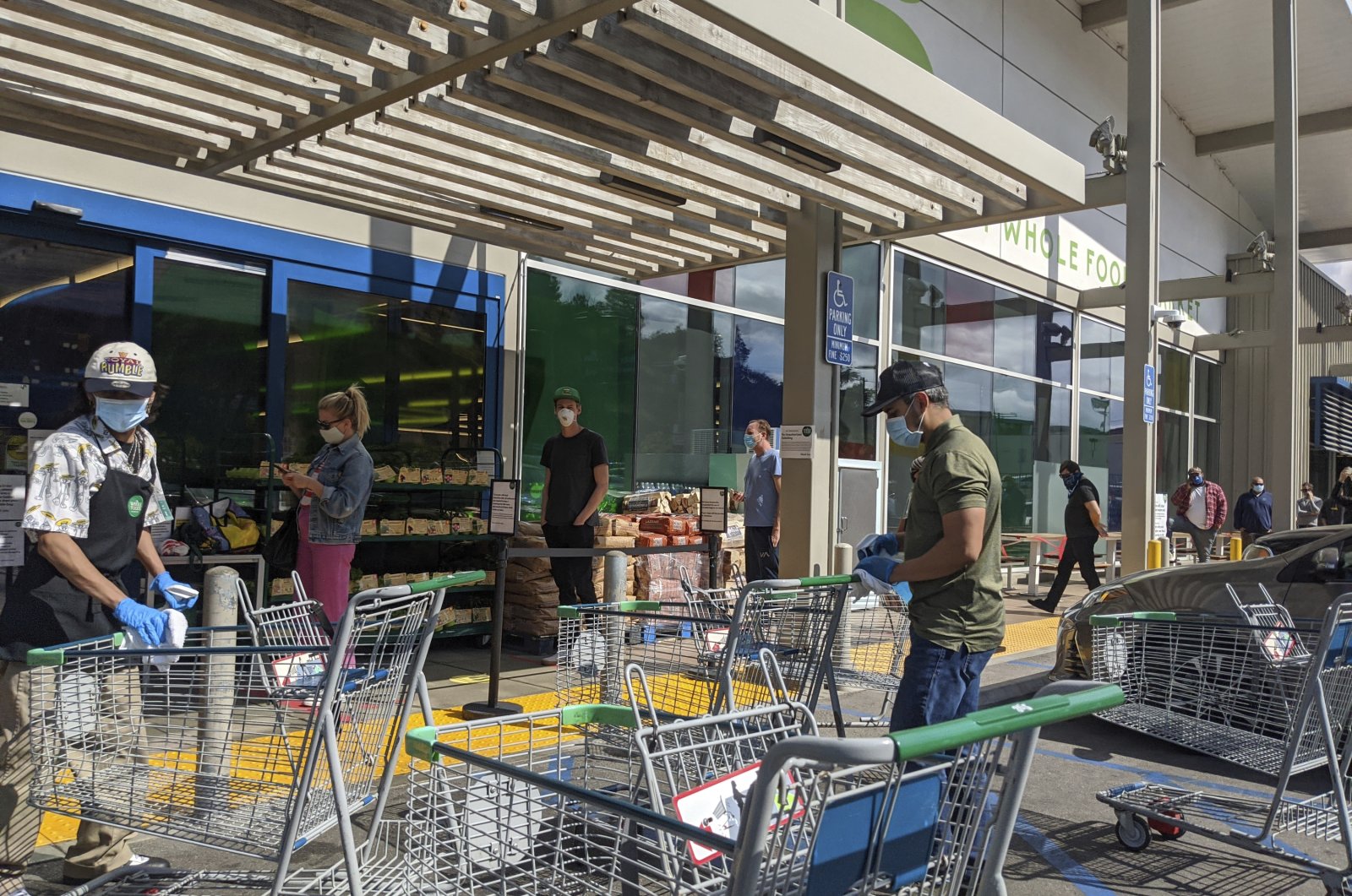Whole Foods Market 365 supermarket workers wear masks and gloves as they sanitize shopping carts, while shoppers wait in line to enter the store, observing social distancing during the coronavirus pandemic, Los Angeles, the U.S., April 17, 2020. (AP File Photo)