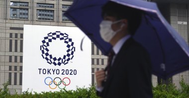 A man wearing a protective mask to help curb the spread of the coronavirus walks in the rain past a banner of the Tokyo 2020 Olympic Games, Tokyo, Japan, May 13, 2021. (AP Photo)