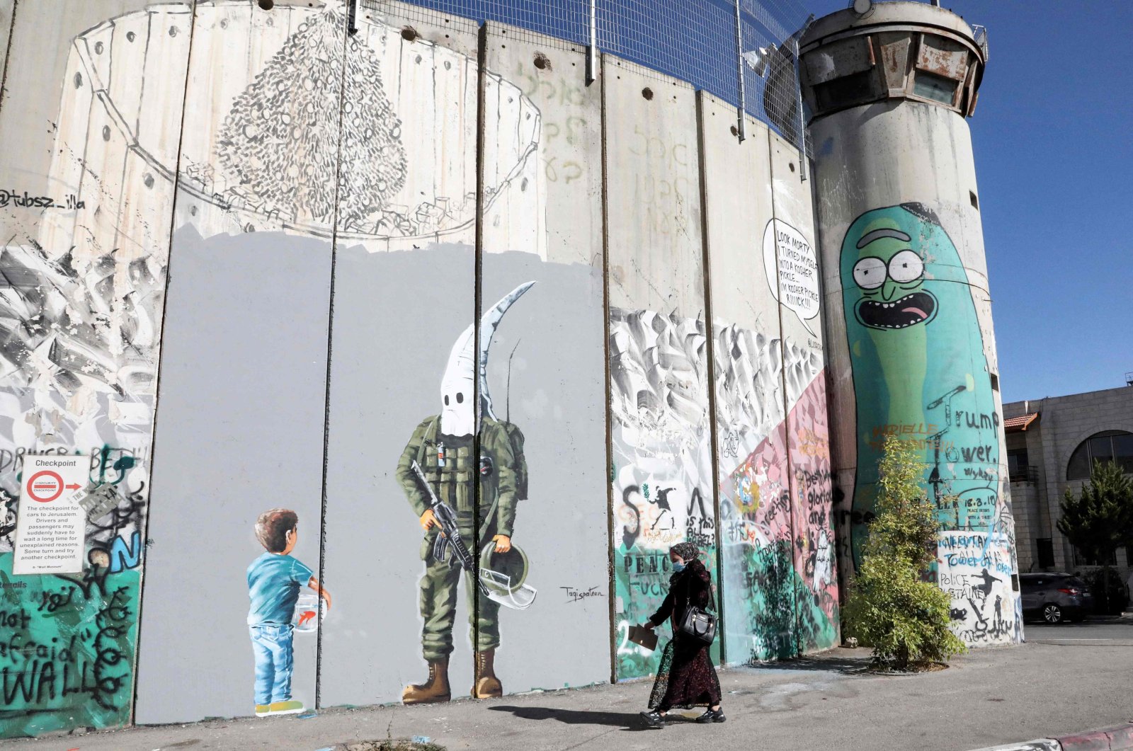 A woman walks past graffiti depicting a boy holding a fishbowl looking at a uniformed Israeli soldier wearing a white hood in the style of the Ku Klux Klan (KKK), drawn by a Palestinian artist along Israel's controversial separation barrier in the city of Bethlehem in the occupied West Bank, Palestine, May 30, 2021. (AFP Photo)
