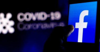 The Facebook logo is seen displayed on a smartphone with a computer model of the COVID-19 coronavirus in the background, April 5, 2020. (Photo by Shutterstock)