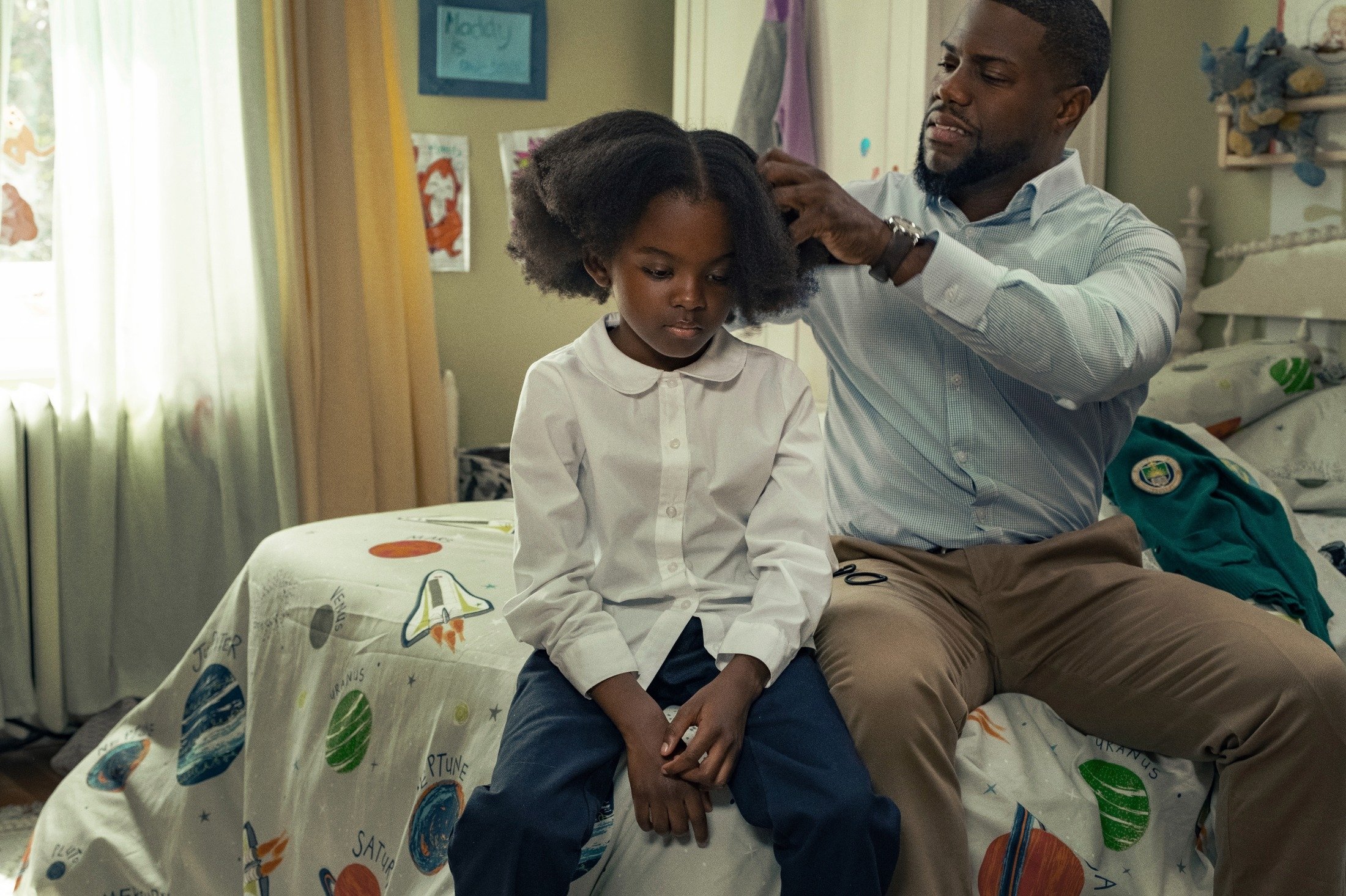Melody Hurd (L) sits on a bed next to Kevin Hart who curbs her hair, in a scene from the movie "Fatherhood." (Netflix via AP)
