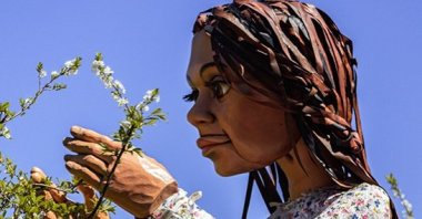 The puppet Little Amal is photographed with branches of a tree. 