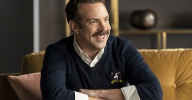 Jason Sudeikis, as Ted Lasso, smiles as he sits on a sofa, in a scene from the series "Ted Lasso." (Apple TV+ via AP)
