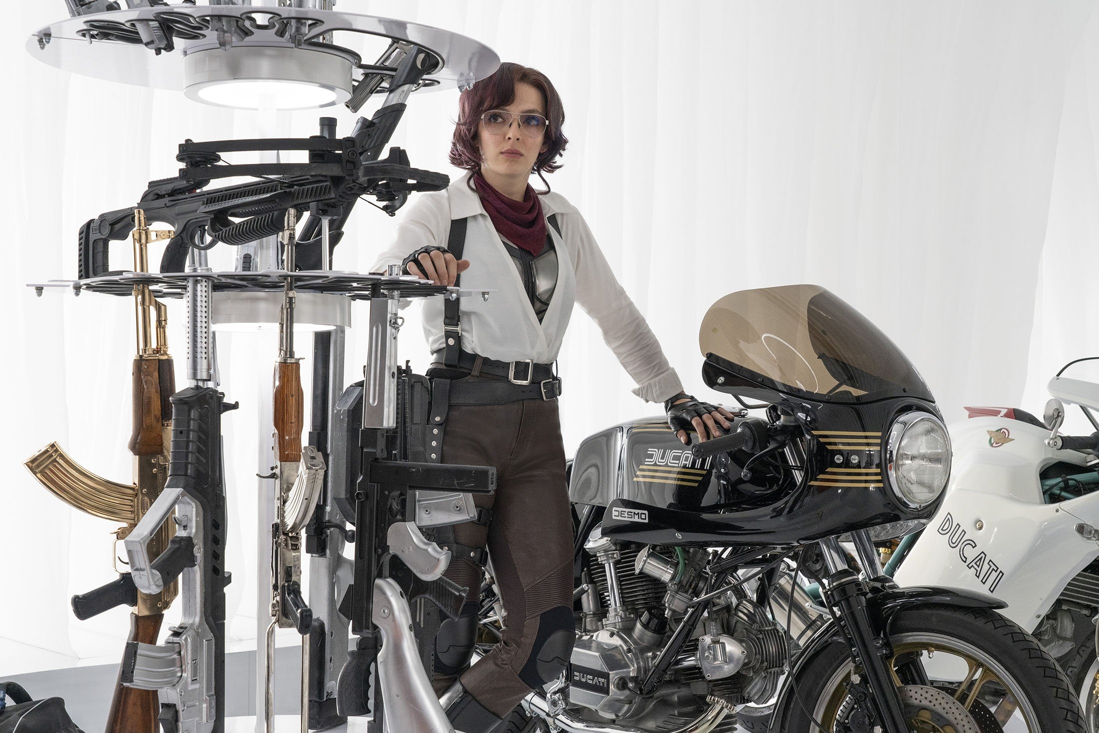 Jodie Comer leans against a motorcycle surrounded by guns in a warehouse of a movie scene 
