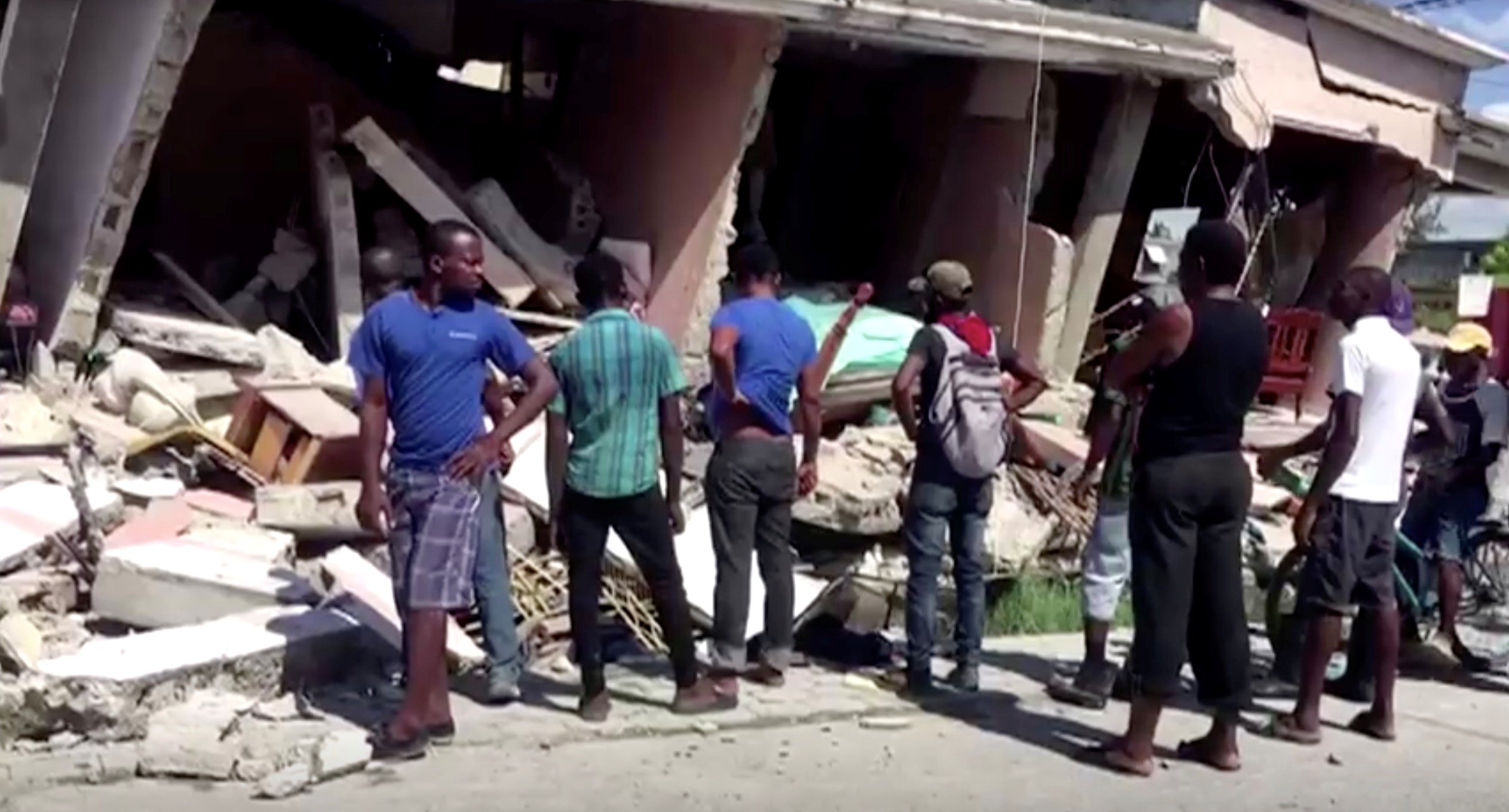 People stand in front of a collapsed building following an earthquake, in Les Cayes, Haiti, in this still image taken from a video obtained by Reuters on August 14, 2021.   (Reuters TV via Reuters)
