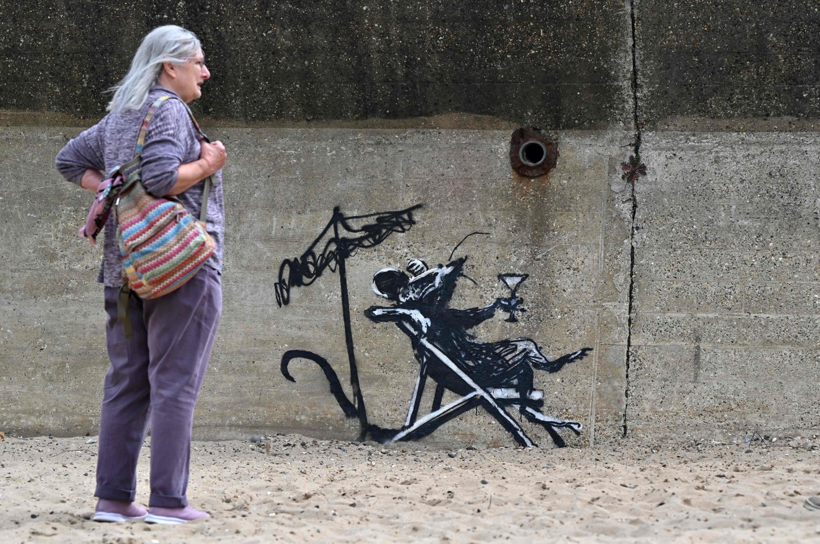 A woman stands beside a graffiti artwork of a rat drinking a cocktail by artist Banksy on a wall at North Beach, Lowestoft, England, Aug. 8, 2021. (AFP Photo)
