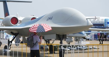 A man carrying an umbrella with the United States flag colors walks past the CH-6 drone during the 13th China International Aviation and Aerospace Exhibition, also known as Airshow China 2021, in Zhuhai in southern China's Guangdong province, Sept. 28, 2021. (AP Photo)