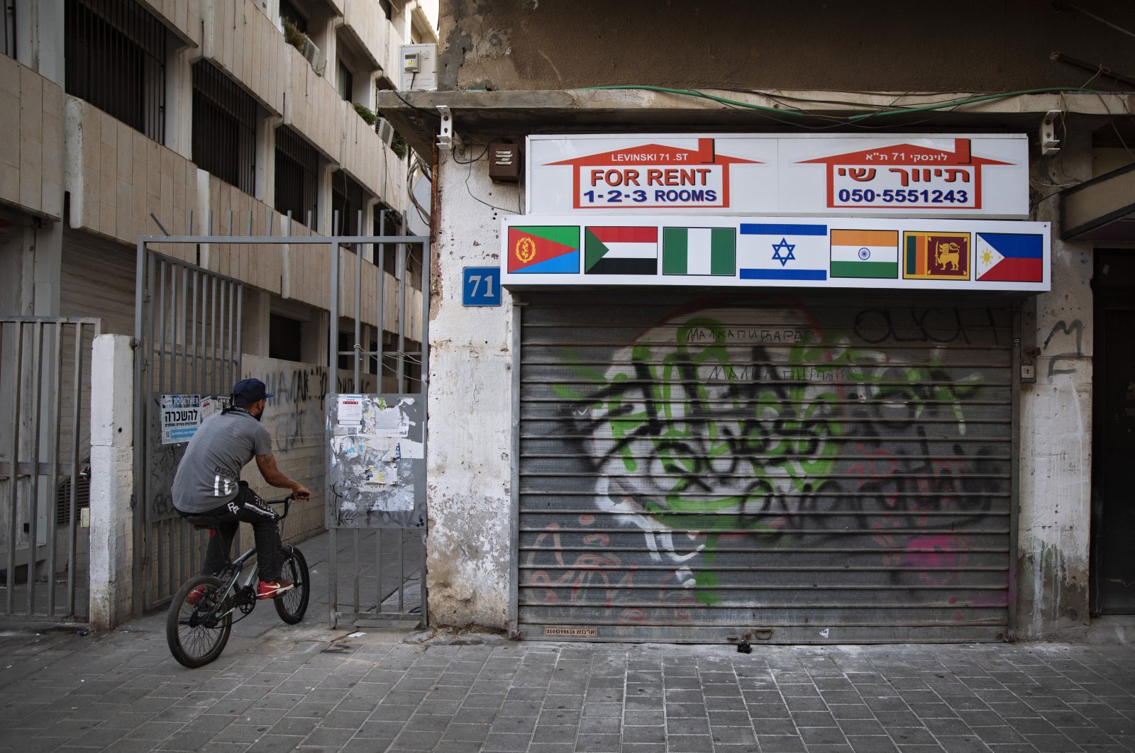 An African migrant rides his bicycle next to a closed real-estate office with the flags of Israel, Sudan and other African countries, in Tel Aviv, Israel, Oct. 23, 2020. (AP Photo)