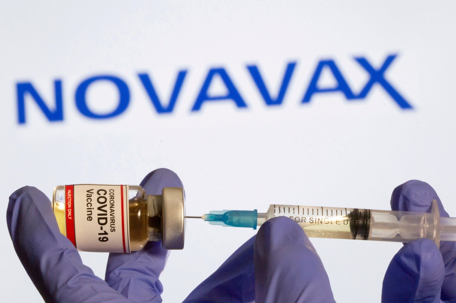 A woman holds a small bottle labeled with a &quot;Coronavirus COVID-19 Vaccine&quot; sticker and a medical syringe in front of displayed Novavax logo in this illustration taken, Oct. 30, 2020. (Reuters Photo)