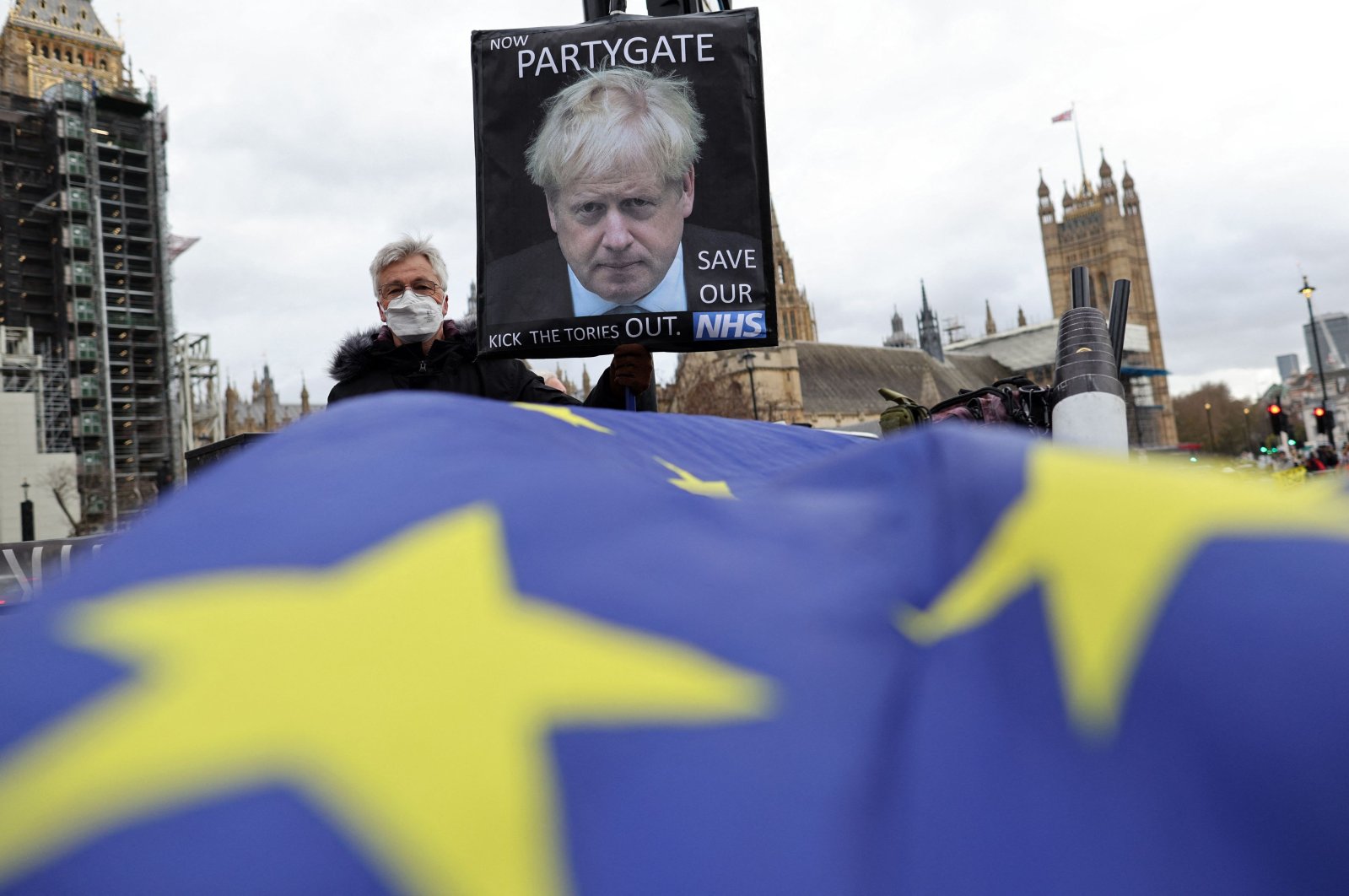 An EU flag flies as an anti-Conservative party protestor holds a placard depicting an image of the party&#039;s leader, and Britain&#039;s Prime Minister, Boris Johnson, reading &quot;now Party-Gate,&quot; as they demonstrate outside of the House of Commons in central London, Dec. 8, 2021. (AFP Photo)