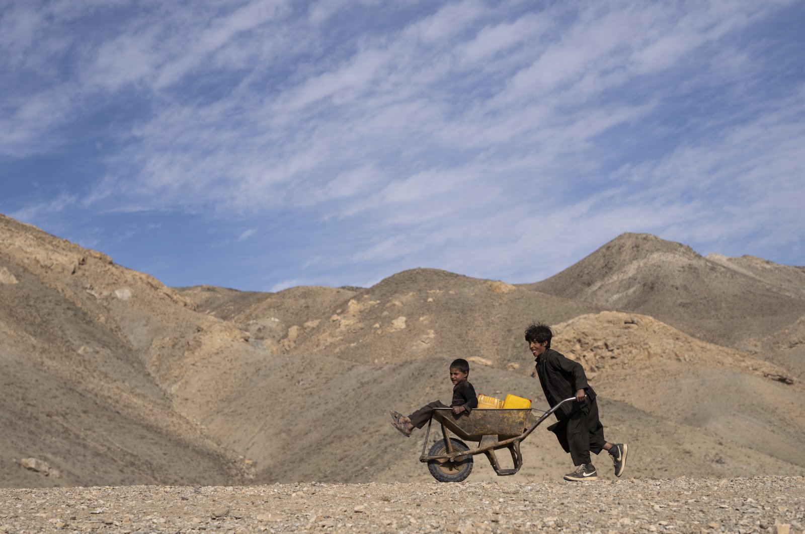 A boy pushes a wheelbarrow with canisters and his younger brother, on their way to collect water from a stagnant pool, about 3 kilometers (2 miles) from their home in Kamar Kalagh village outside Herat, Afghanistan, Nov. 26, 2021. (AP Photo)