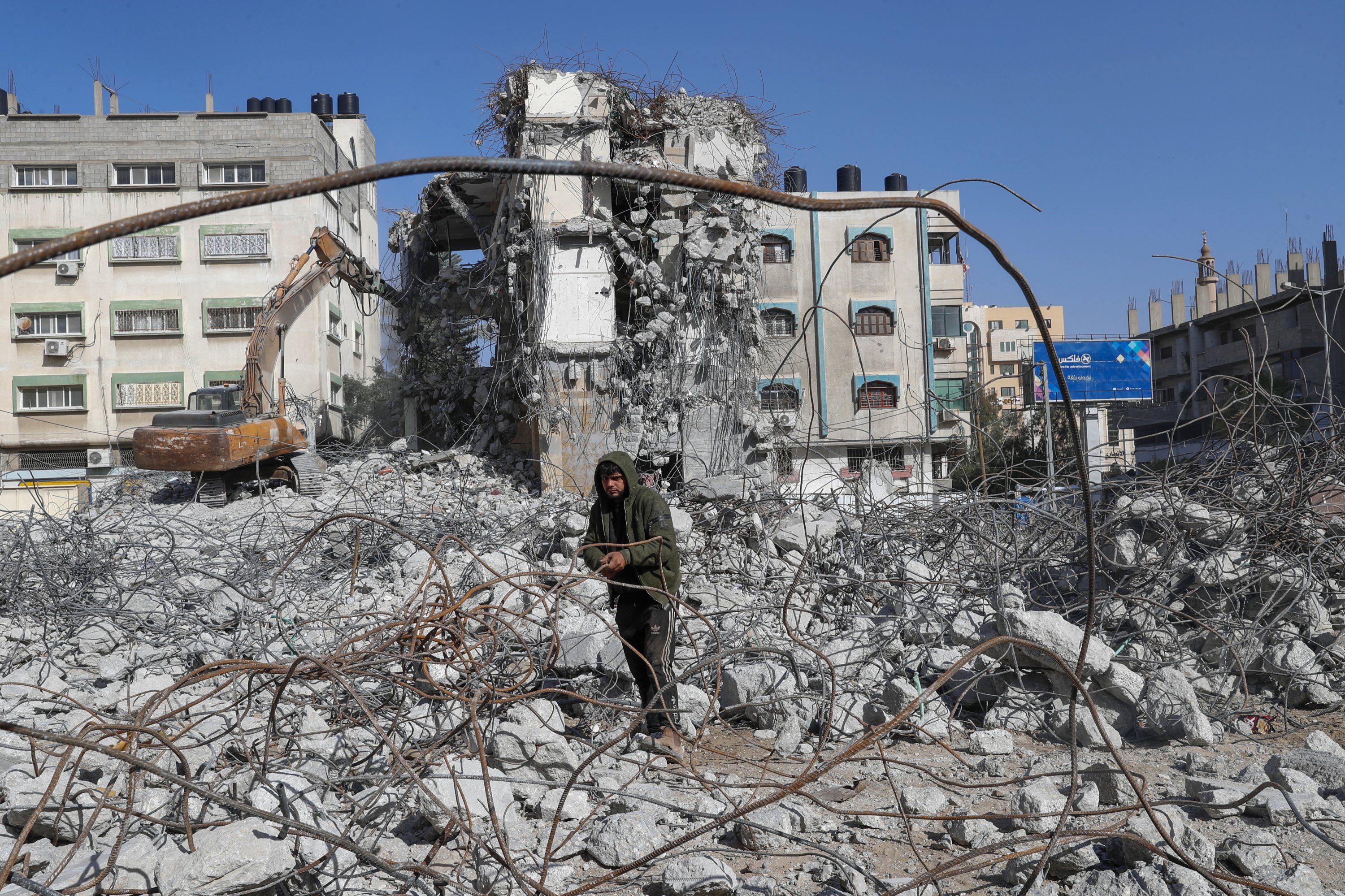 A backhoe breaks and remove parts of the Al-Jawhara building, as a worker recycles metal iron rods from the rubble of the building, which was damaged by Israeli airstrikes during Israel's war with Gaza's Hamas rulers last May, in the central of al-Rimal neighborhood of Gaza City, Jan. 10, 2022. (AP Photo)