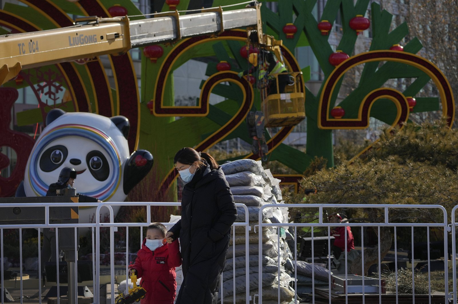 A woman and a child wearing face masks to help protect them from the coronavirus walk by workers setting up decorations for the Winter Olympic Games and Lunar New Year in Beijing, China, Jan. 16, 2022. (AP Photo)