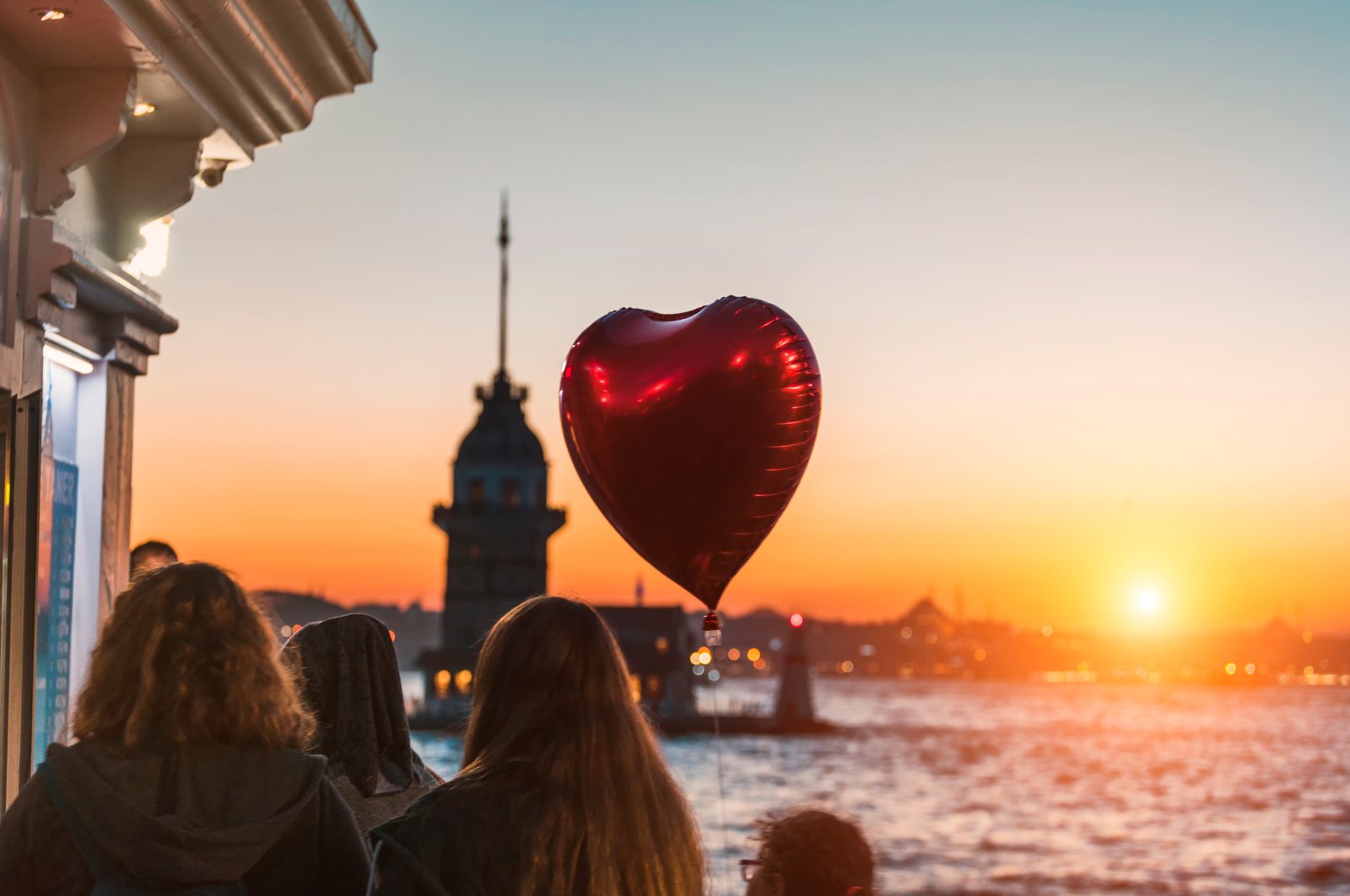 A red heart-shaped balloon flies over the heads of girls enjoying an amazing view of the Maiden&#039;s Tower at sunset, Istanbul, Turkey. (Shutterstock Photo)