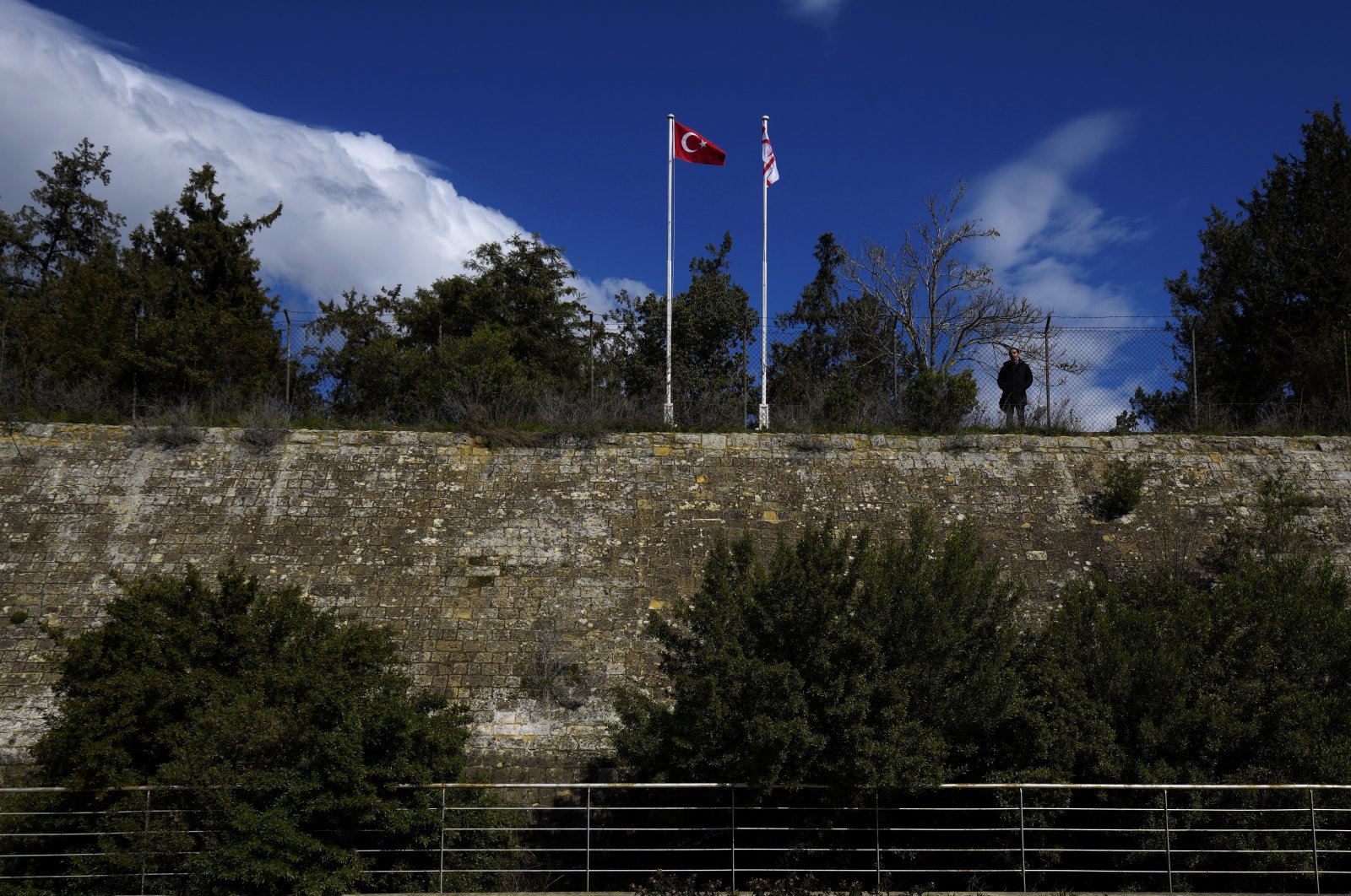 A man stands behind a fence and looks from the Turkish side of the island to the Greek side in the south next to Turkish and Turkish Cypriot flags in the capital Lefkoşa (Nicosia), TRNC, Feb. 10, 2022. (AP Photo)