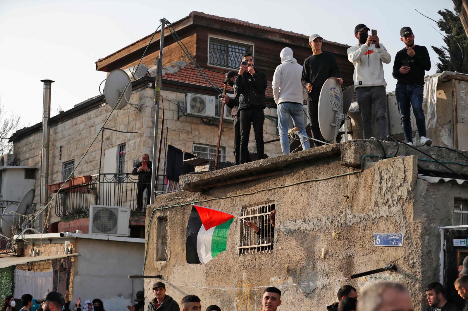 Palestinian demonstrators gather atop the roof of one of the houses facing eviction, while one waves a Palestinian flag from a window below, during a protest in the flashpoint neighbourhood of Sheikh Jarrah in Israeli-annexed east Jerusalem on February 18, 2022. - Hundreds of Palestinians face eviction by Israel from homes in Sheikh Jarrah and other east Jerusalem neighbourhoods. Circumstances surrounding the eviction threats vary. (Photo by Ahmad GHARABLI / AFP)