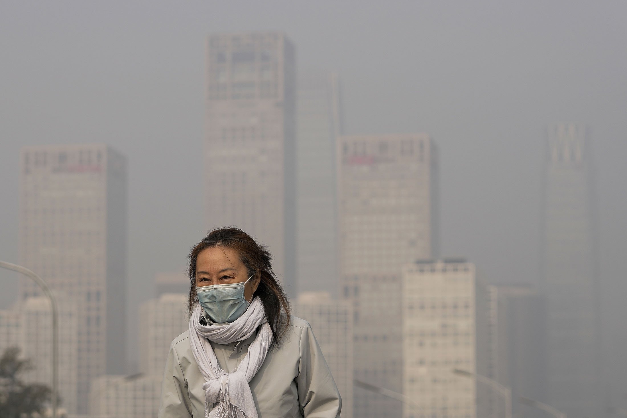 Seorang wanita mengenakan masker wajah untuk melindungi diri dari COVID-19 berjalan di depan gedung perkantoran di Kawasan Pusat Bisnis yang diselimuti kabut polusi di Beijing, Cina, 18 November 2021. (AP Photo)