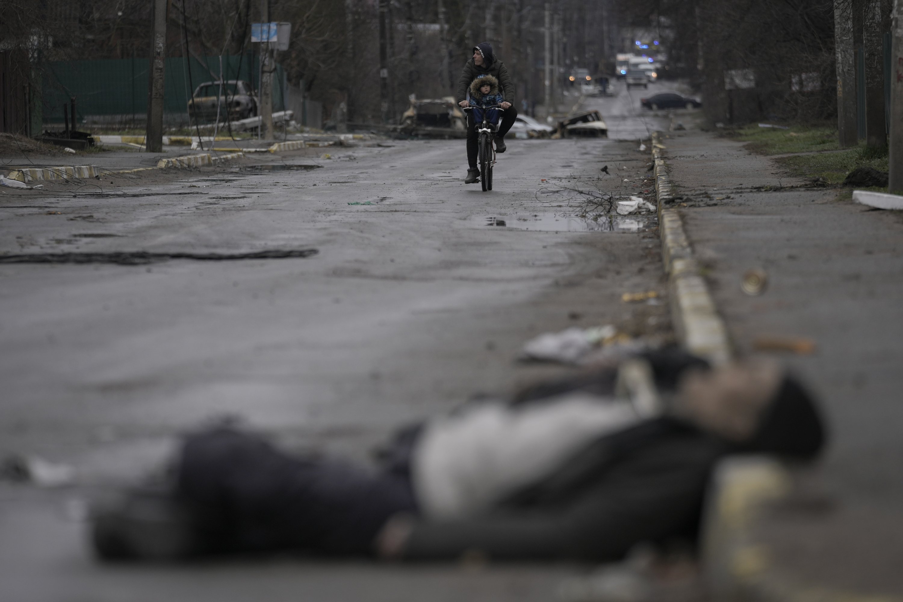 Seorang pria dan anak-anak mengendarai sepeda saat mayat warga sipil tergeletak di jalan di pinggiran kota Bucha, Ukraina, yang sebelumnya diduduki Rusia, 2 April 2022. (AP Photo)