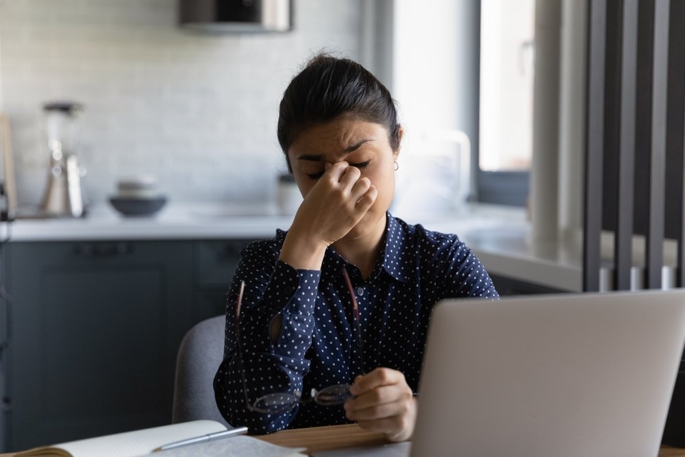 Exhausted young woman working on computer takes off glasses possibly suffering from migraine or headache. (Shutterstock Photo)