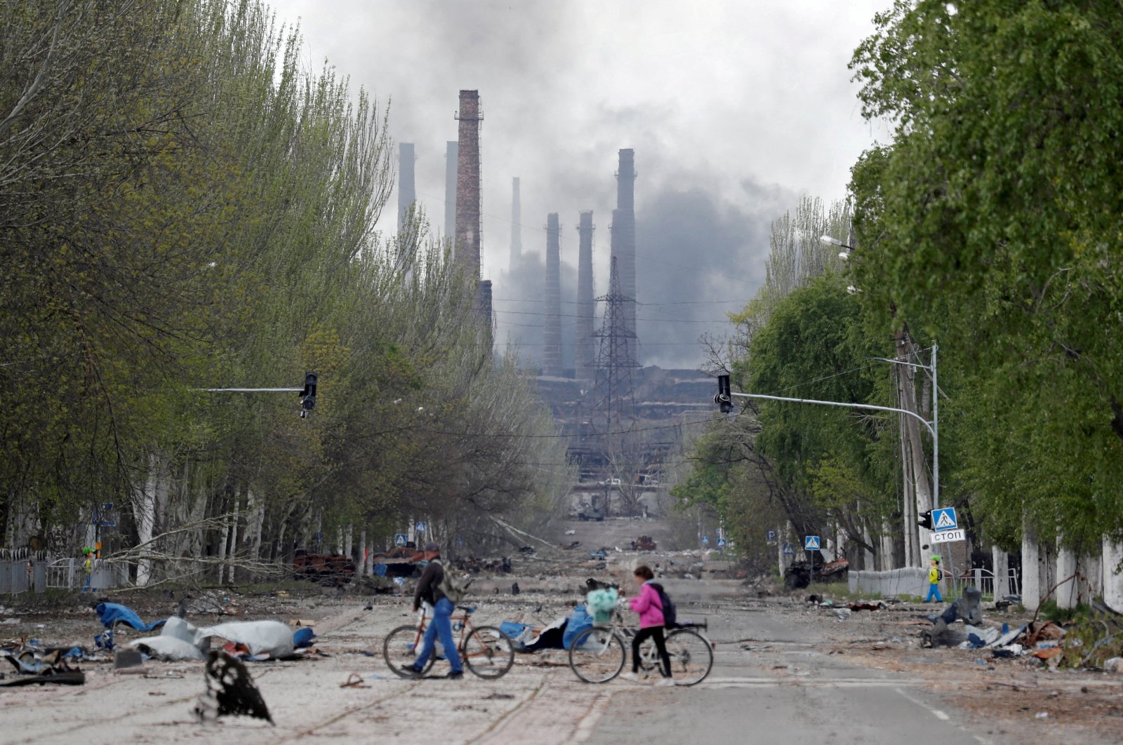 People walk their bikes across the street as smoke rises above a plant of Azovstal Iron and Steel Works during the Russian invasion of Ukraine, in the southern port city of Mariupol, Ukraine, May 2, 2022. (Reuters Photo)