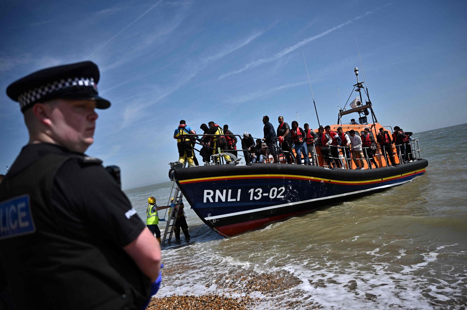 A British police officer stands guard on the beach of Dungeness, on the southeast coast of England, on June 15, 2022, as Royal National Lifeboat Institution&#039;s (RNLI) members of staff help migrants to disembark from one of their lifeboat after they were picked up at sea while attempting to cross the English Channel. (AFP Photo)