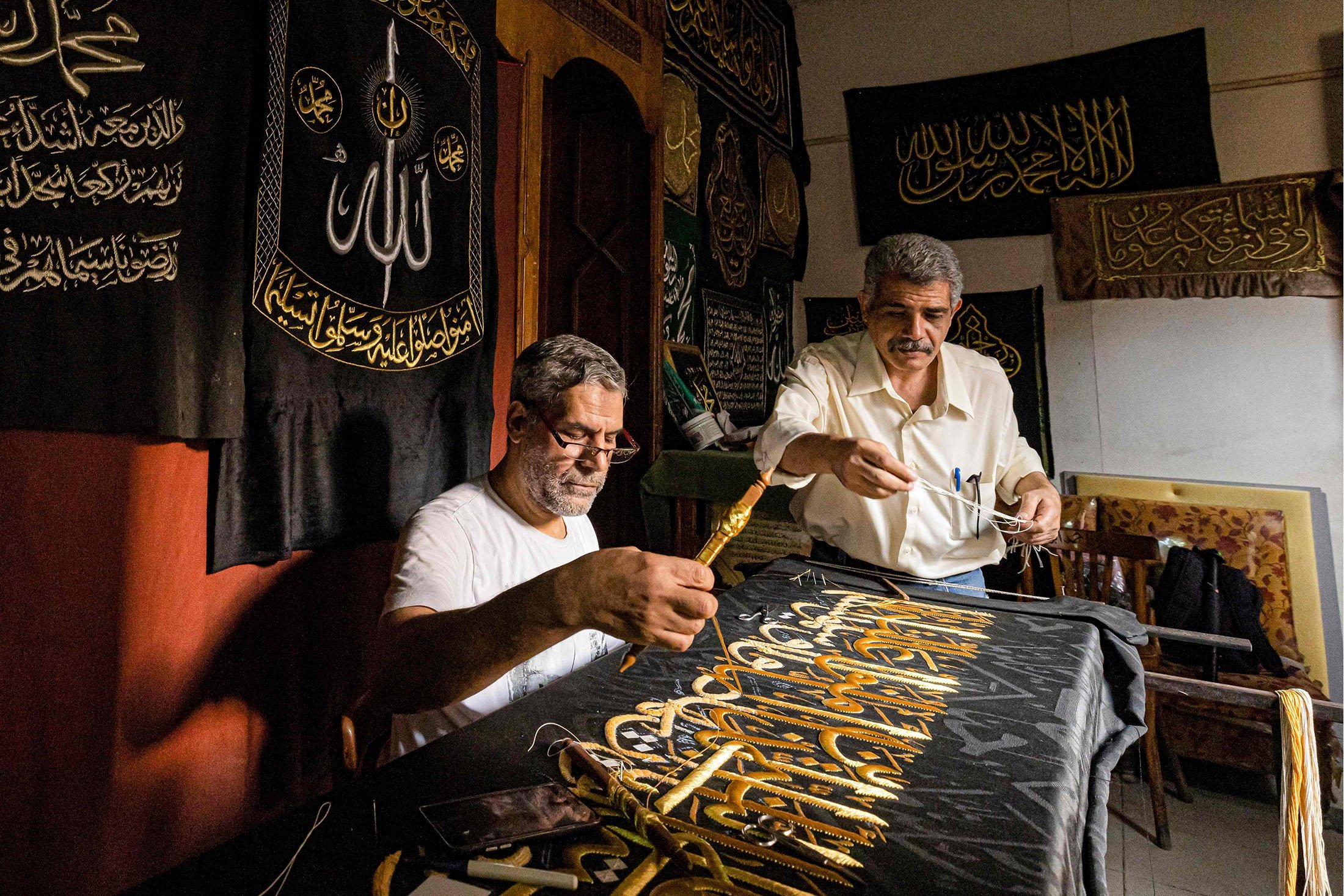 Penjahit Mesir Ahmed Othman el-Kassabgy (kanan) mengawasi saat karyawan lain (kiri) menjahit dengan benang emas sebuah ayat dari Al-Qur'an ke tirai replika untuk dijual sebagai suvenir bagi wisatawan yang berkunjung ke Kairo, Mesir, 15 Juni 2022. (AFP Foto)