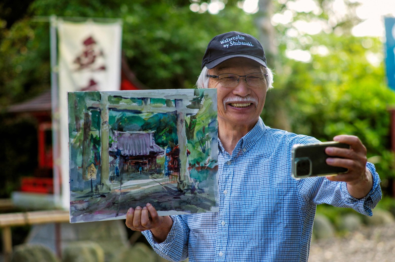 Japanese art instructor Harumichi Shibasaki poses with his finished watercolor painting while holding up his phone at a shrine in Isumi, Chiba prefecture, Japan, May 25, 2022. (AFP)
