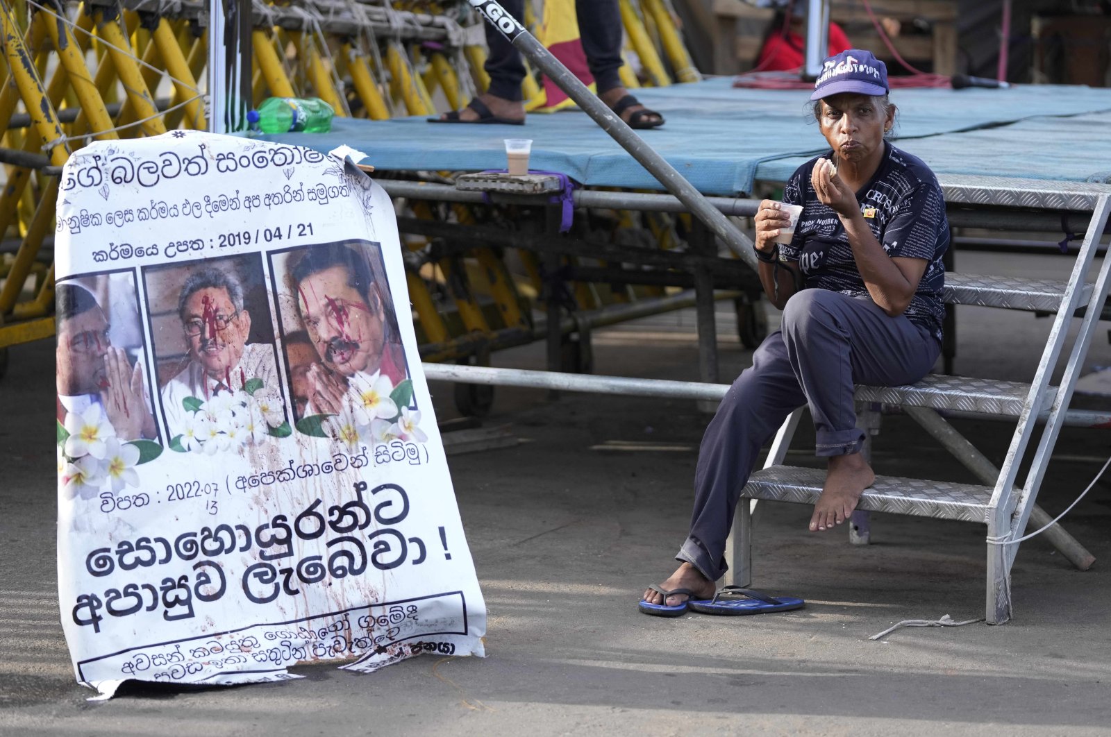 A protester drinks a cup of tea as she sits by a defaced poster carrying portraits of ousted President Gotabaya Rajapaksa (C) and his brothers at the entrance to the president&#039;s office in Colombo, Sri Lanka, July 15, 2022. (AP Photo)