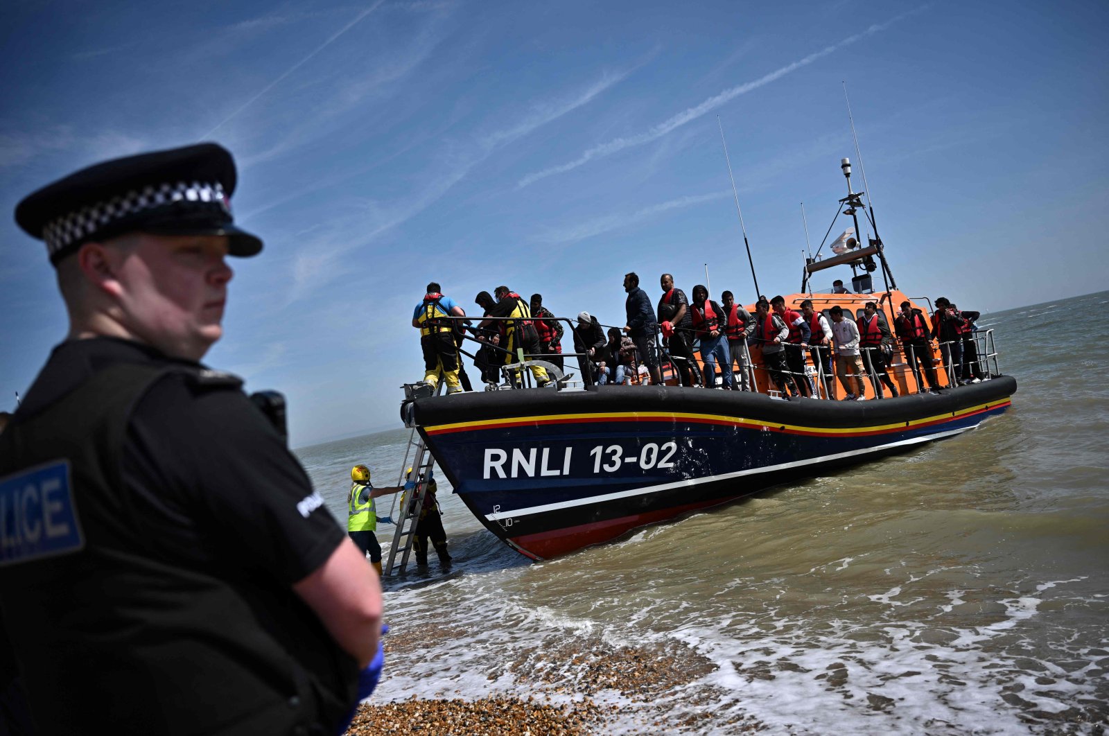 A British police officer stands guard on the beach of Dungeness, on the southeast coast of England, as Royal National Lifeboat Institution&#039;s (RNLI) members help migrants to disembark from one of their lifeboats after they were picked up at sea while attempting to cross the English Channel, June 15, 2022. (AFP Photo)