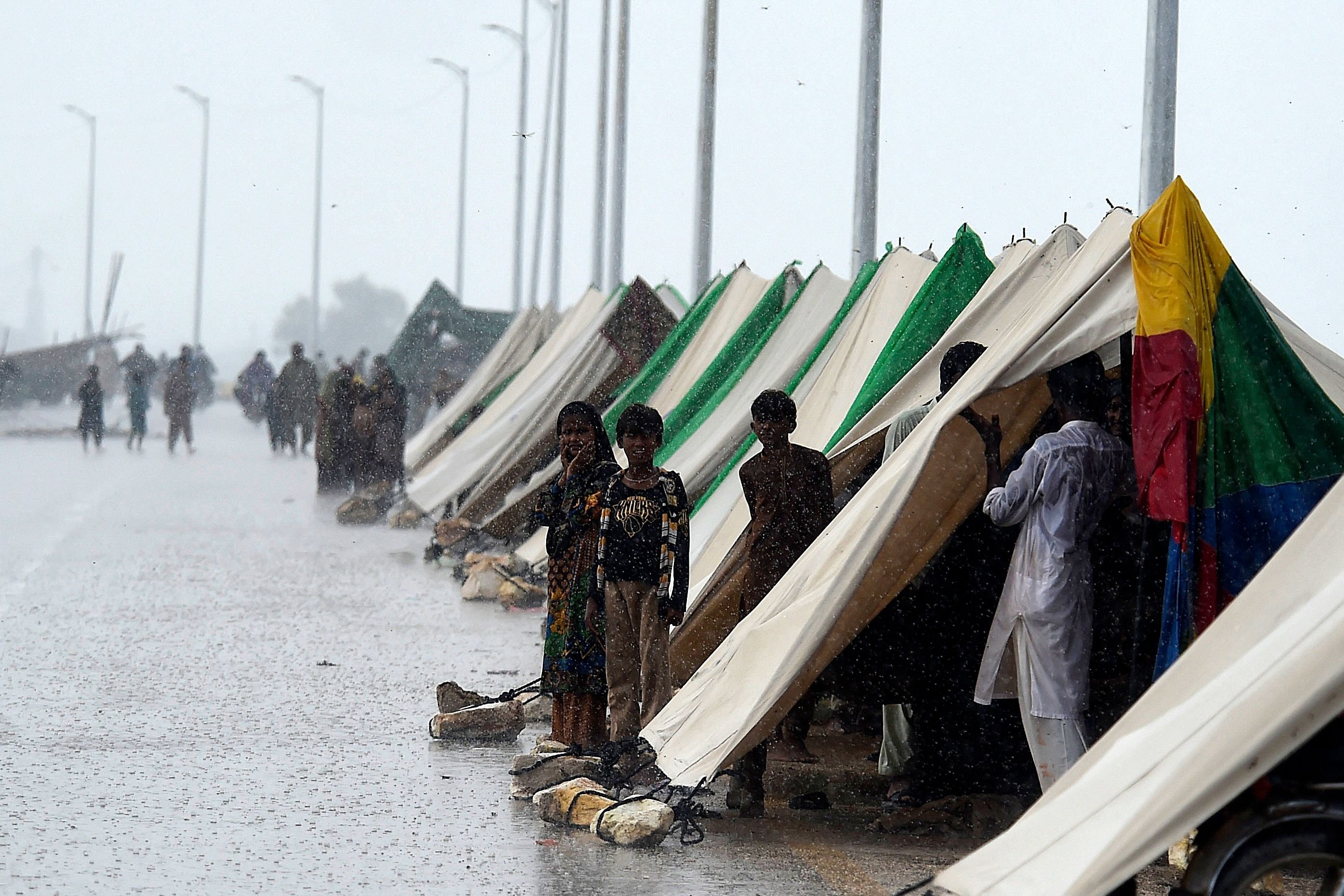 Orang-orang berdiri di luar tenda sementara yang didirikan di sepanjang jalan saat hujan deras di Sukkur, Sindh, Pakistan, 27 Agustus 2022. (AFP Photo)
