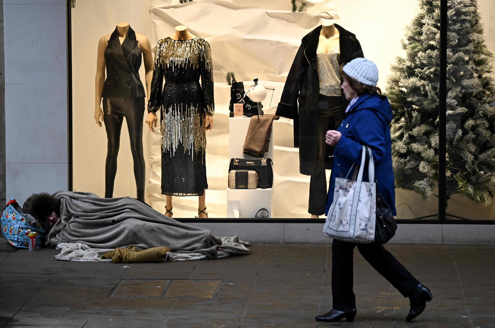 A pedestrian walks past a homeless person sleeping on the floor in front of a clothing shop decorated for Christmas in Chester, U.K., Nov. 17, 2022. (AFP Photo)