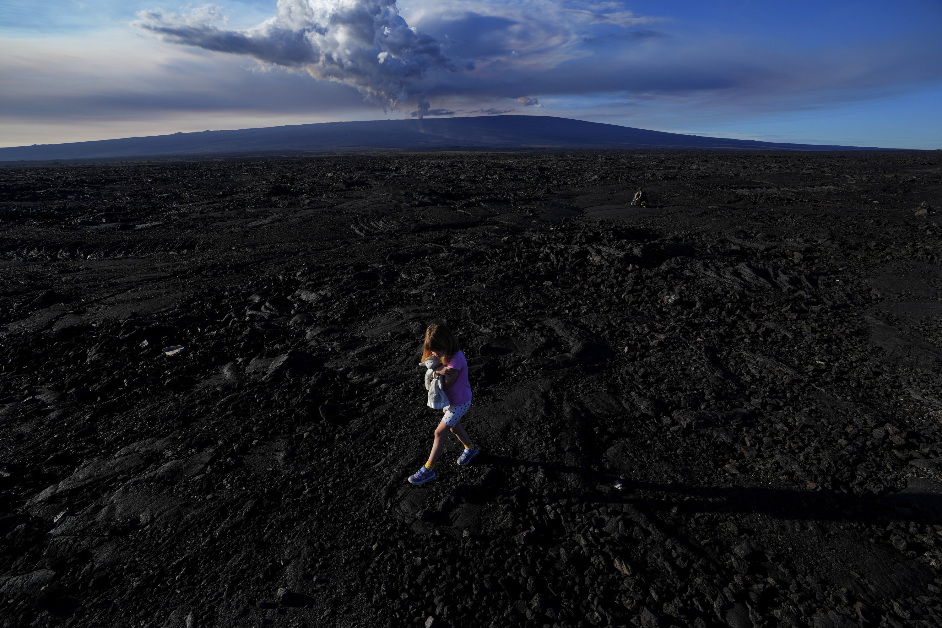Abigail Dewar, dari Alberta, Kanada, memegang boneka binatang saat berjalan di atas batu lava yang mengeras dari letusan sebelumnya saat gunung berapi Mauna Loa meletus, dekat Hilo, Hawaii, AS, 30 November 2022. (Foto AP)