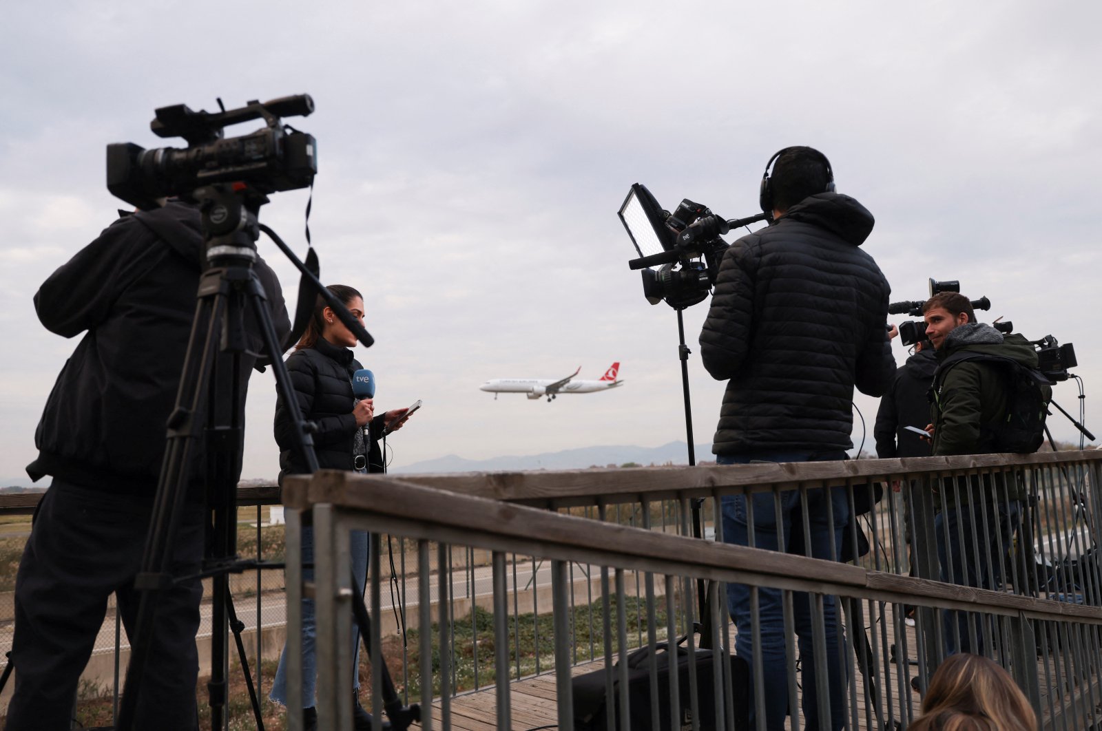 A plane flies in the background as journalists work, after a commercial airplane flying from Morocco to Türkiye made an emergency landing in Barcelona&#039;s El Prat airport in the early hours and 28 would-be migrants on board ran away across the tarmac in Barcelona, Spain Dec. 7, 2022. (Reuters Photo)