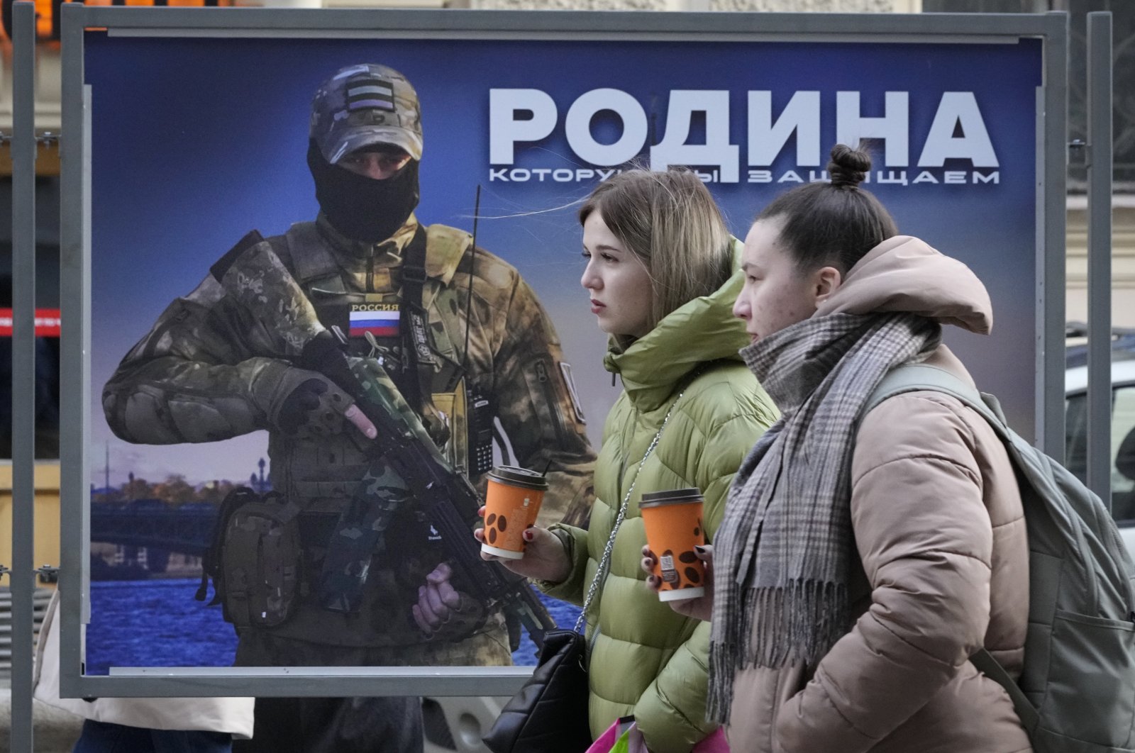 Girls walk past a stand with an image of a Russian serviceman and words &#039;The Motherland we defend&#039; at a street exhibition of military photos in St. Petersburg, Russia, Feb. 9, 2023. (AP Photo)